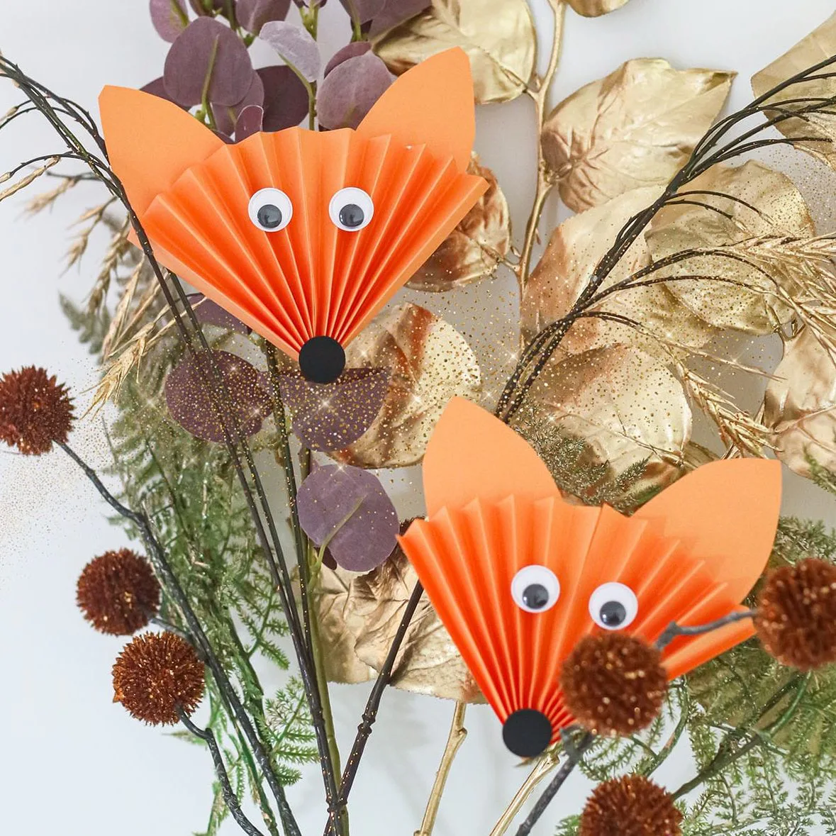 Zwei gefaltete orange Papierfüchse mit Wackelaugen zwischen Herbstdeko