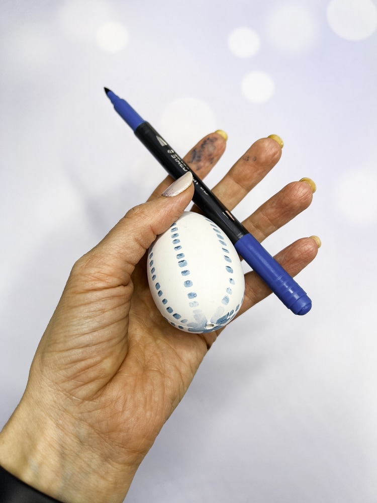 Hand hält ein mit blauem Stift bemaltes Plastikei aus dem Malset