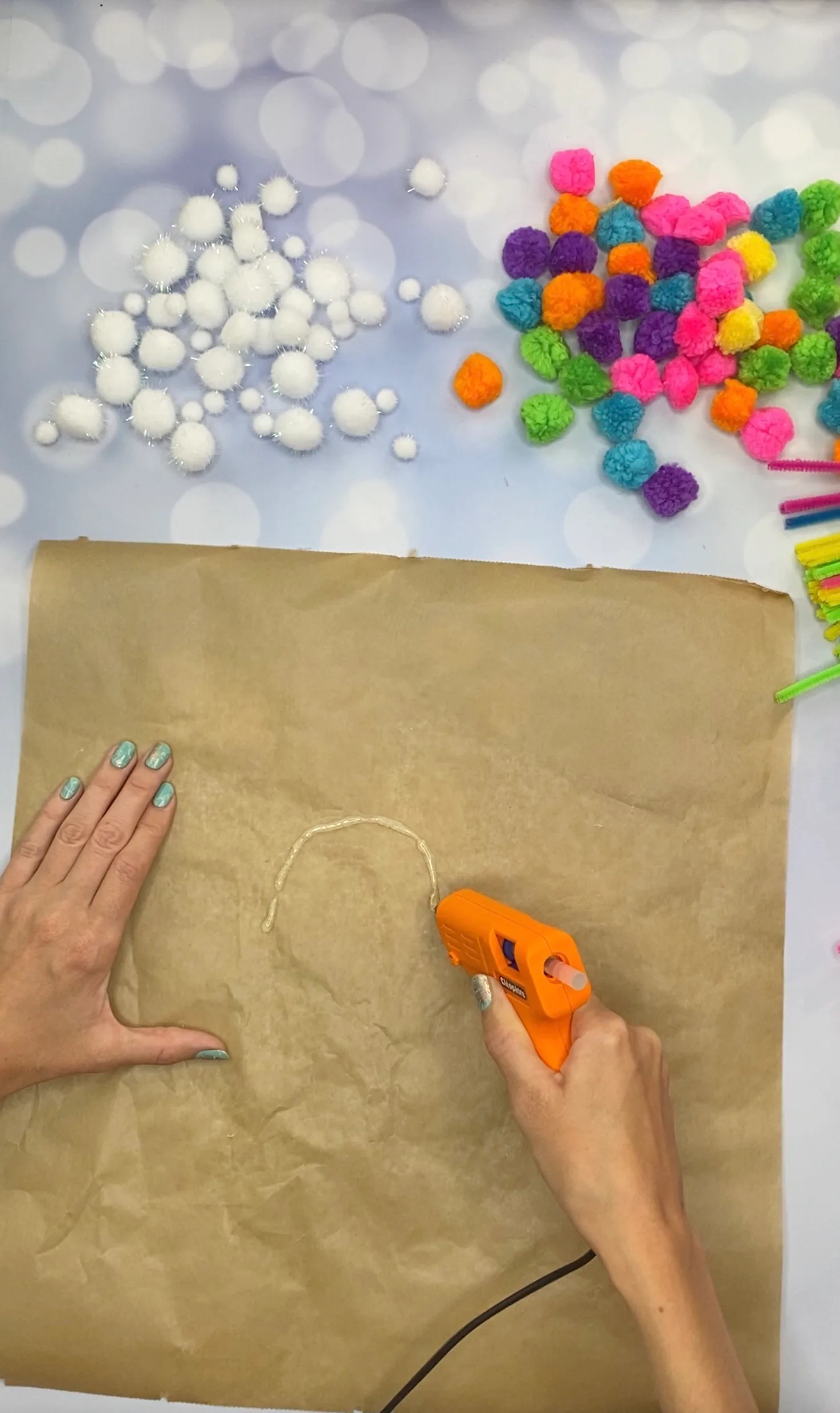 Heißkleber wird in Bogenform auf Backpapier aufgetragen, daneben liegen bunte Pompons bereit